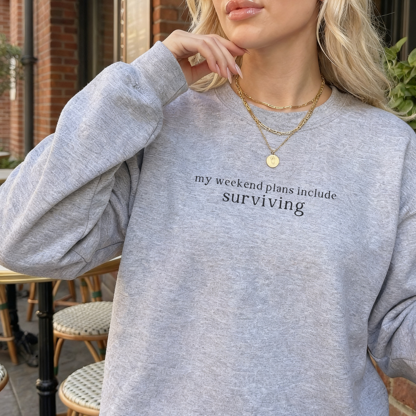 a woman wearing a sport grey sweatshirt with text 'my weekend plans include surviving' styled with gold necklaces at a cafe
