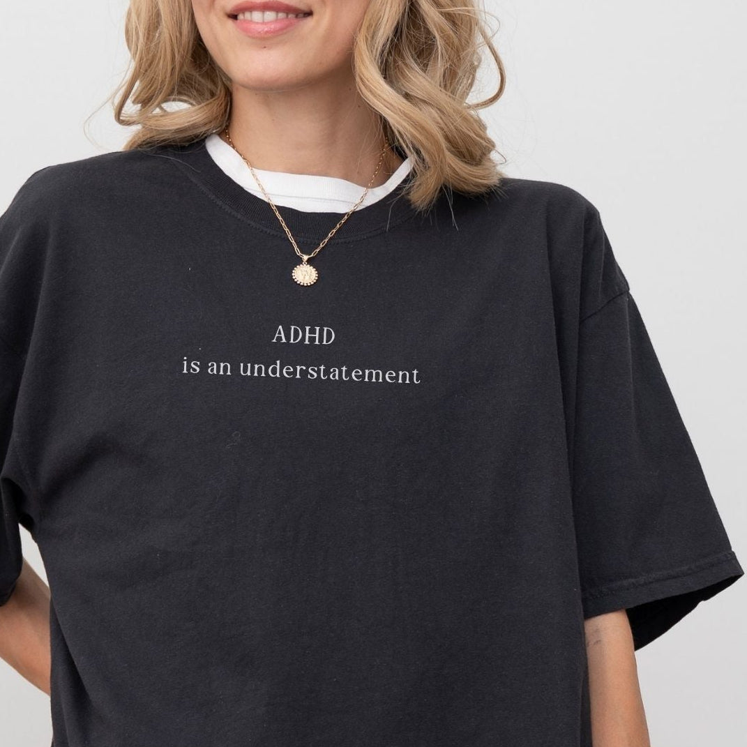 Oversized washed black t-shirt with minimalist white text that reads 'ADHD is an understatement'. Model wears a simple gold chain necklace, showcasing the vintage, aesthetic streetwear fit against a clean white background.