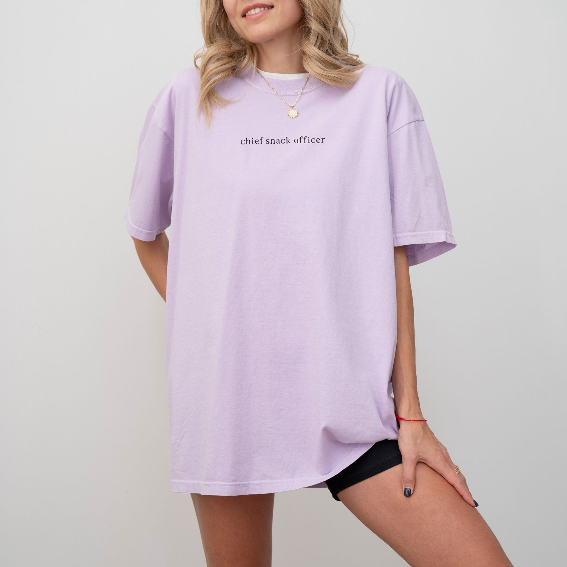 Woman wearing an orchid 'Chief Snack Officer' oversized t-shirt and a gold necklace. Minimalist studio product photography for the Barely Sane clothing collection.