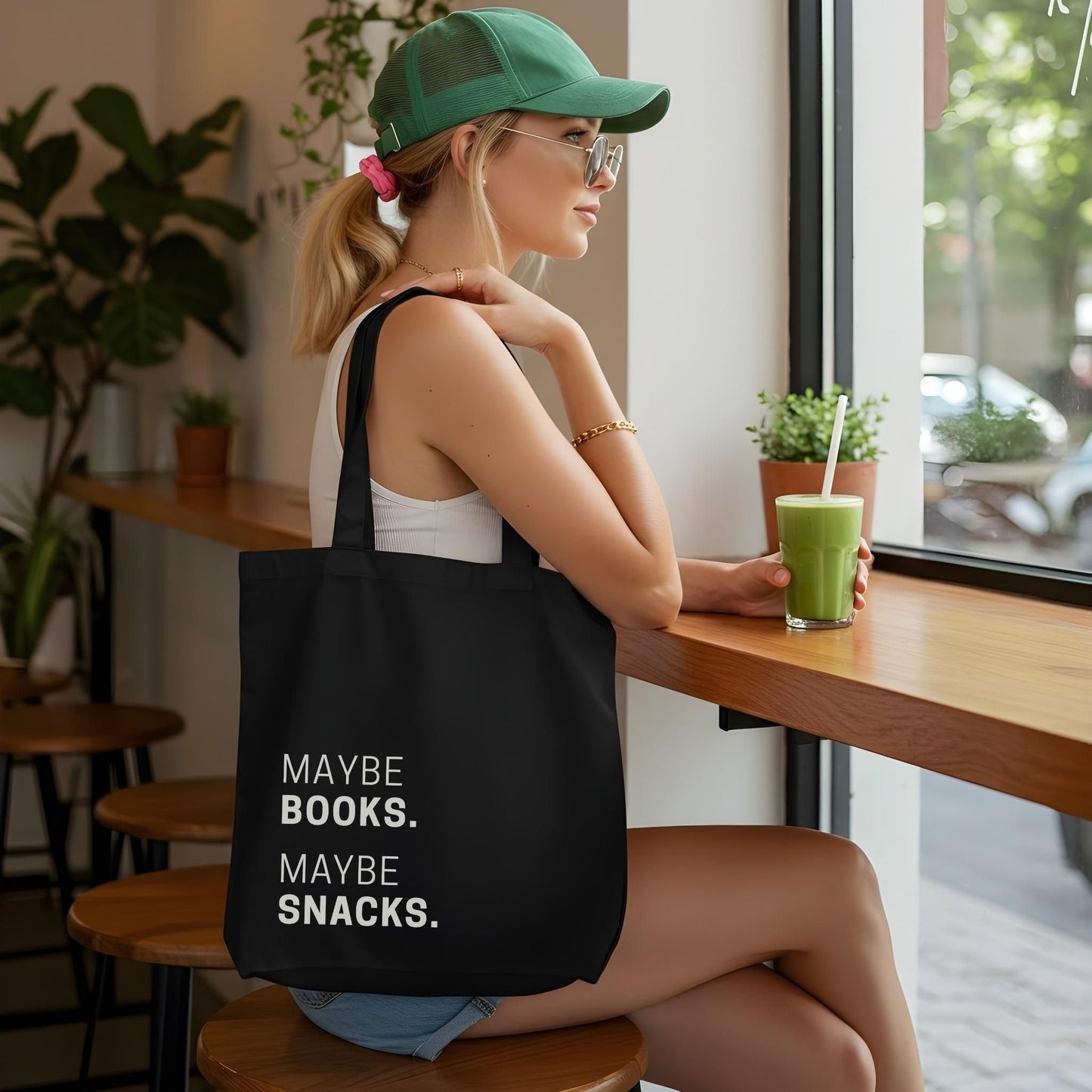 A woman wearing a green baseball cap and sunglasses sitting in a cafe, carrying the black 'Maybe Books. Maybe Snacks.' tote bag. Aesthetic lifestyle product photography for Barely Sane's casual collection.