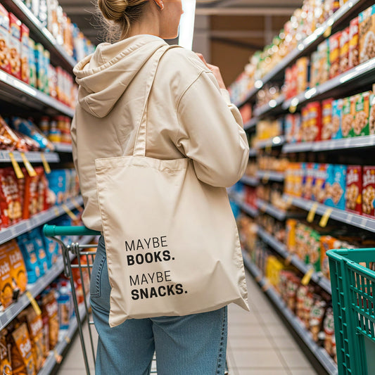 A woman in a beige hoodie holding a shopping cart, carrying the natural cotton 'Maybe Books. Maybe Snacks.' tote bag in a grocery store aisle. Aesthetic lifestyle apparel by Barely Sane.