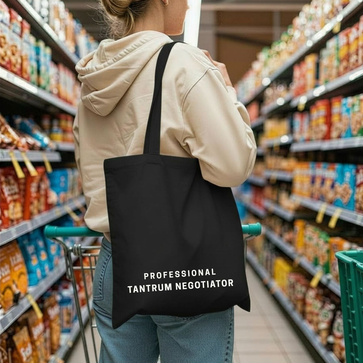 A woman in a beige hoodie holding a shopping cart, carrying the black 'Professional Tantrum Negotiator' tote bag in a grocery store aisle. Aesthetic lifestyle apparel by Barely Sane.