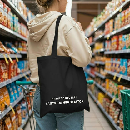 A woman in a beige hoodie holding a shopping cart, carrying the black 'Professional Tantrum Negotiator' tote bag in a grocery store aisle. Aesthetic lifestyle apparel by Barely Sane.