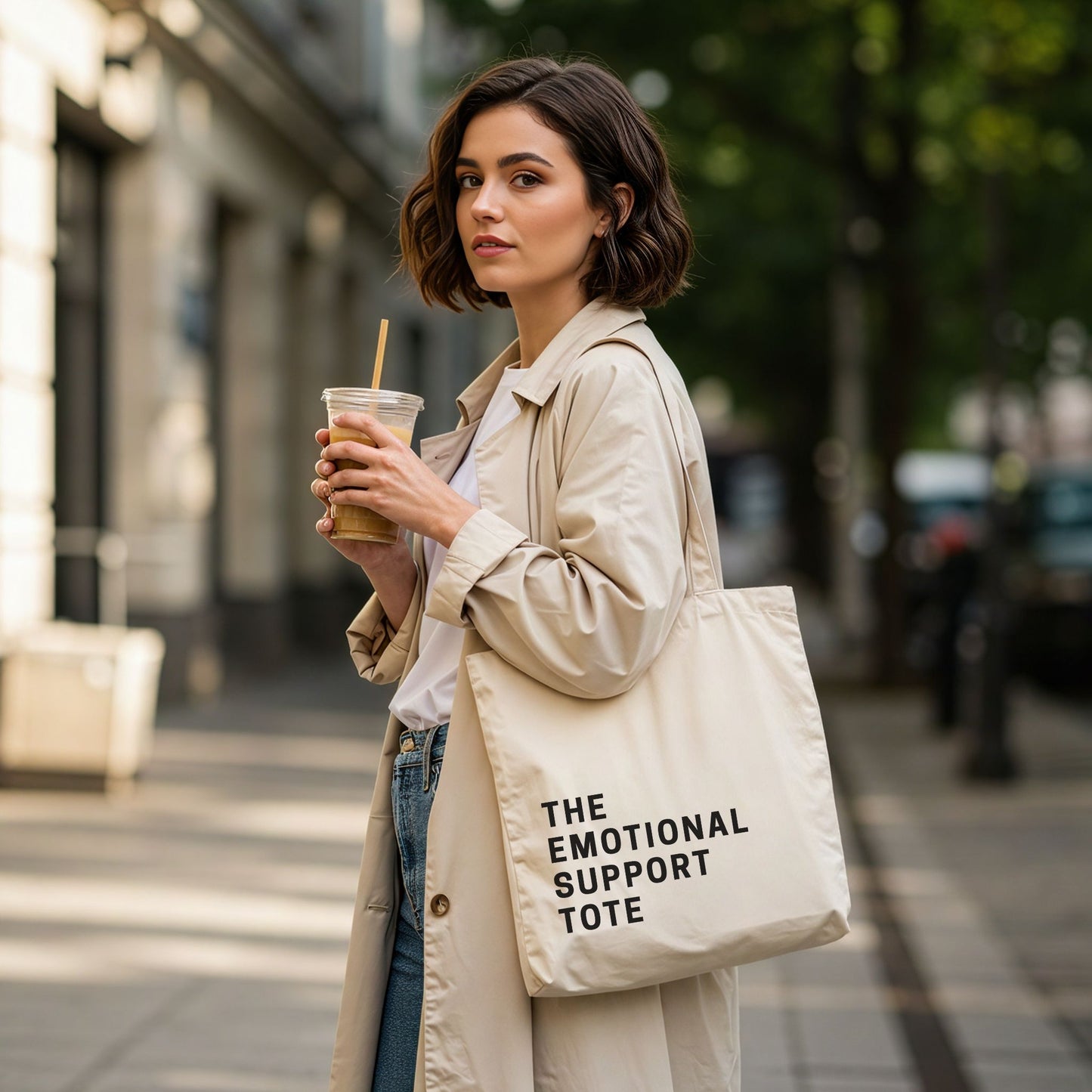 'The Emotional Support Tote' in natural cotton canvas. A woman in a trench coat holds the tote bag and an iced coffee in a city setting. Aesthetic and durable everyday bag by Barely Sane.