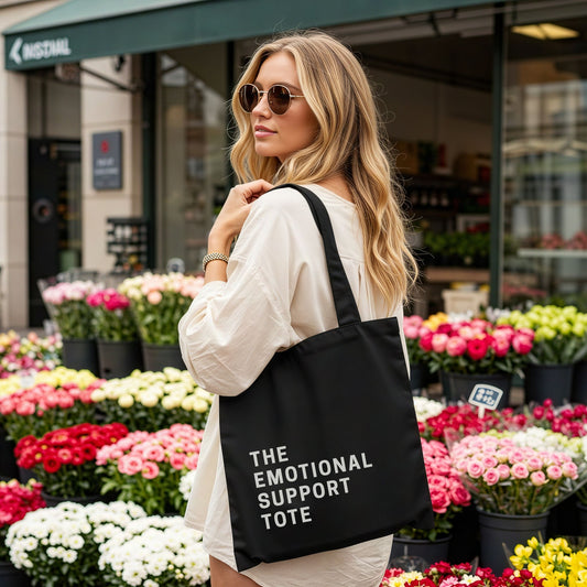 Woman holding a black tote bag labeled 'The Emotional Support Tote' in front of a flower shop.