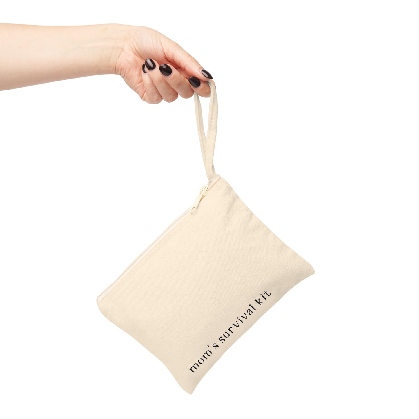 A woman's hand with black nail polish holding a cream canvas zipper pouch by its wristlet strap. The pouch features the text "mom's survival kit" printed on the front against a white background.