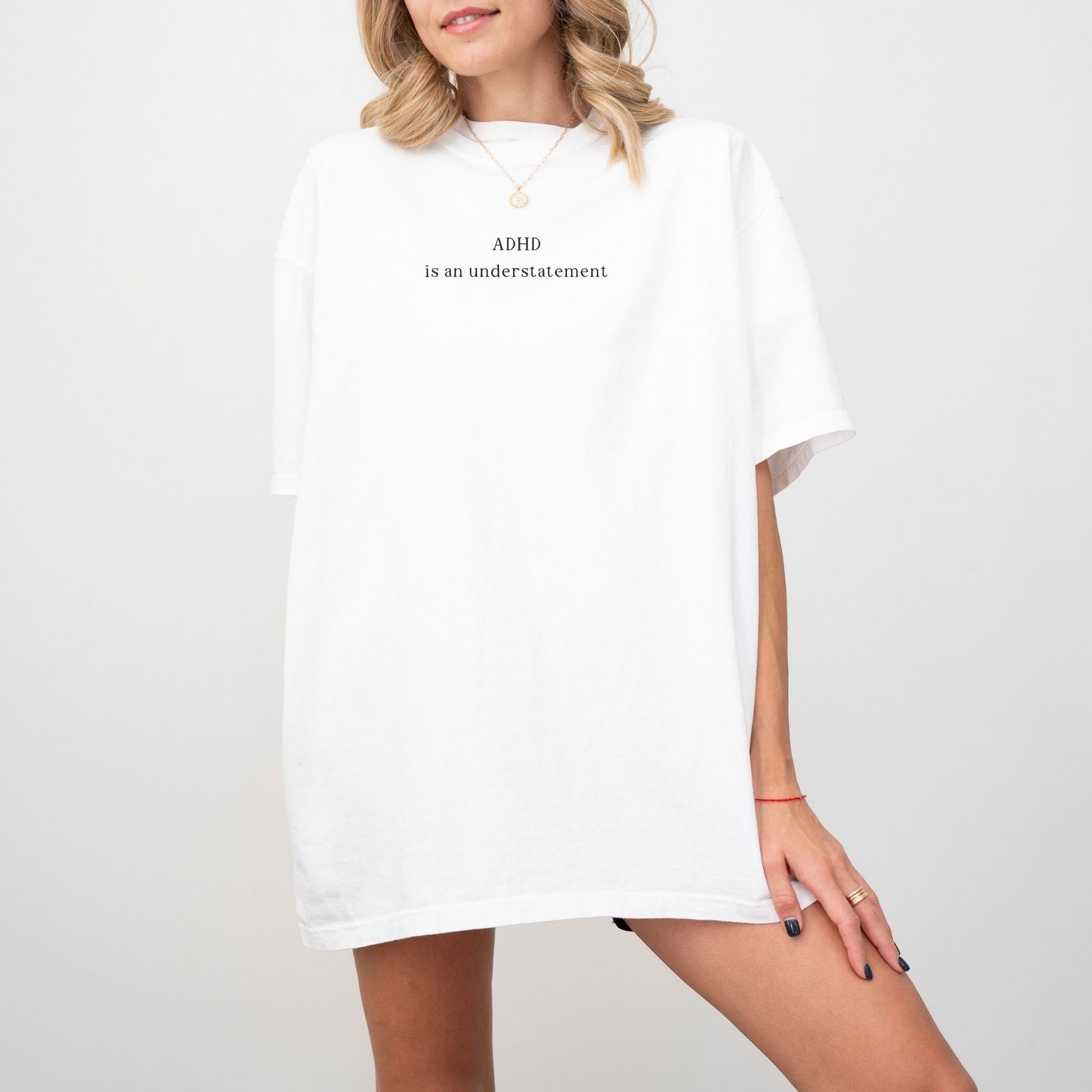 Oversized classic white graphic t-shirt with minimalist black text that reads 'ADHD is an understatement'. Blonde model wears a simple gold chain necklace, showcasing the bright, aesthetic streetwear fit against a clean white background.