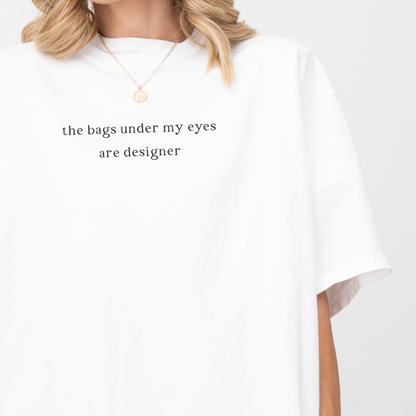 A close-up of a woman wearing a white 'the bags under my eyes are designer' oversized t-shirt. She is accessorized with a gold necklace. Relatable fashion, lifestyle photography for the Barely Sane clothing collection.