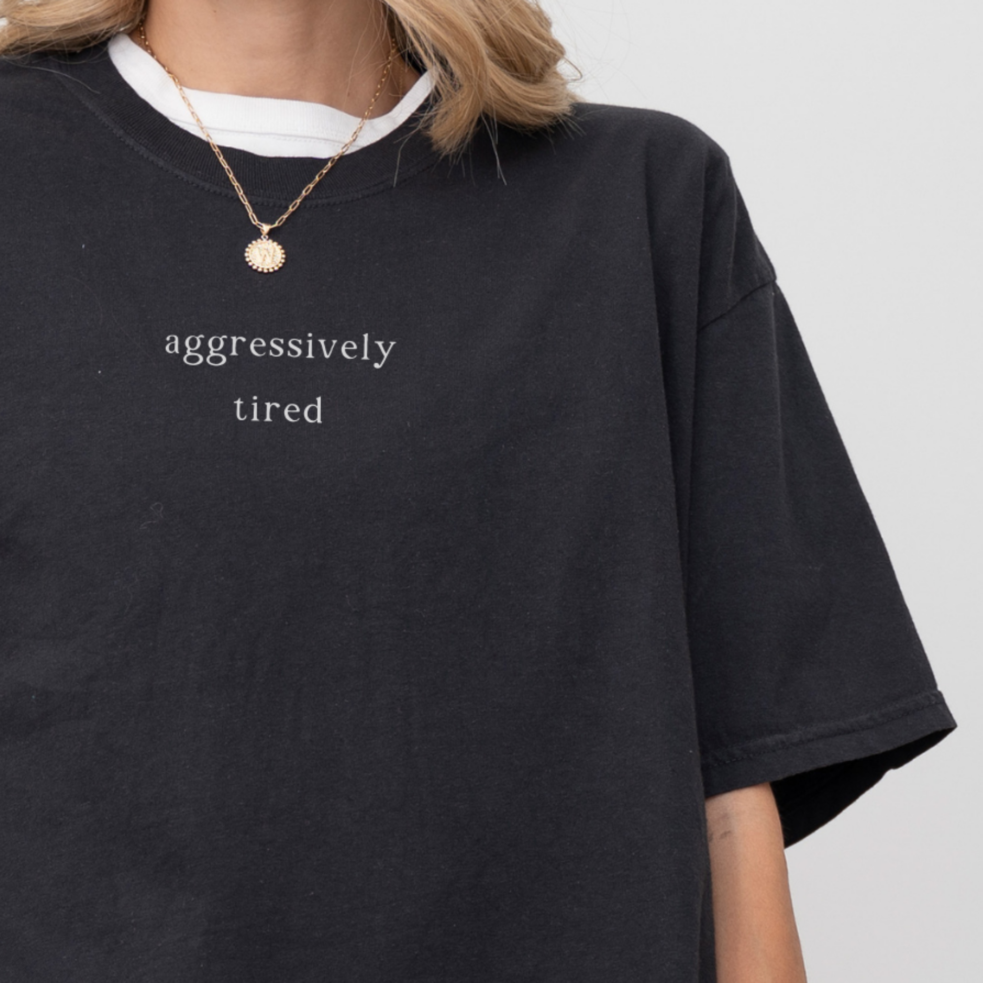 Close up of a black t-shirt with 'aggressively tired' text worn by a person on a plain background