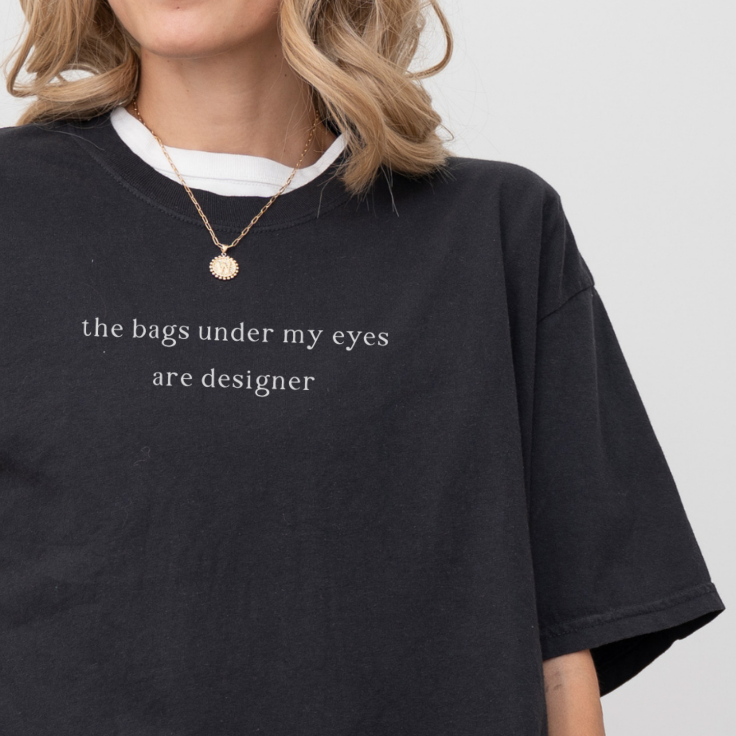 A close-up of a woman wearing a black 'the bags under my eyes are designer' oversized t-shirt. She is accessorized with a gold necklace. Relatable fashion, lifestyle photography for the Barely Sane clothing collection.
