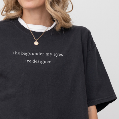 A close-up of a woman wearing a black 'the bags under my eyes are designer' oversized t-shirt. She is accessorized with a gold necklace. Relatable fashion, lifestyle photography for the Barely Sane clothing collection.