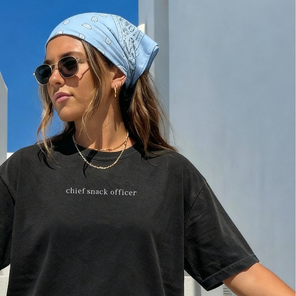 A woman wearing a light blue bandana, sunglasses, and the black 'Chief Snack Officer' t-shirt. Aesthetic and casual summer lifestyle photography for Barely Sane's clothing collection.