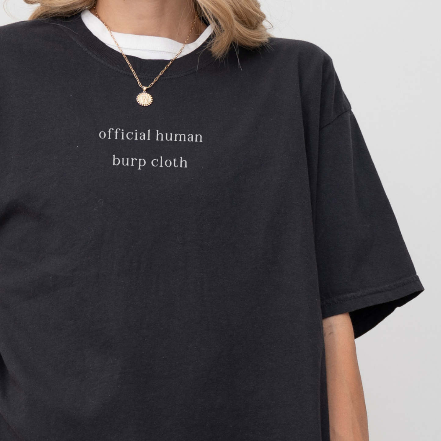A close-up of a woman wearing a black oversized t-shirt with the white text 'official human burp cloth' printed on the chest. She is wearing a gold coin necklace. Minimalist studio product photography for the Barely Sane collection.