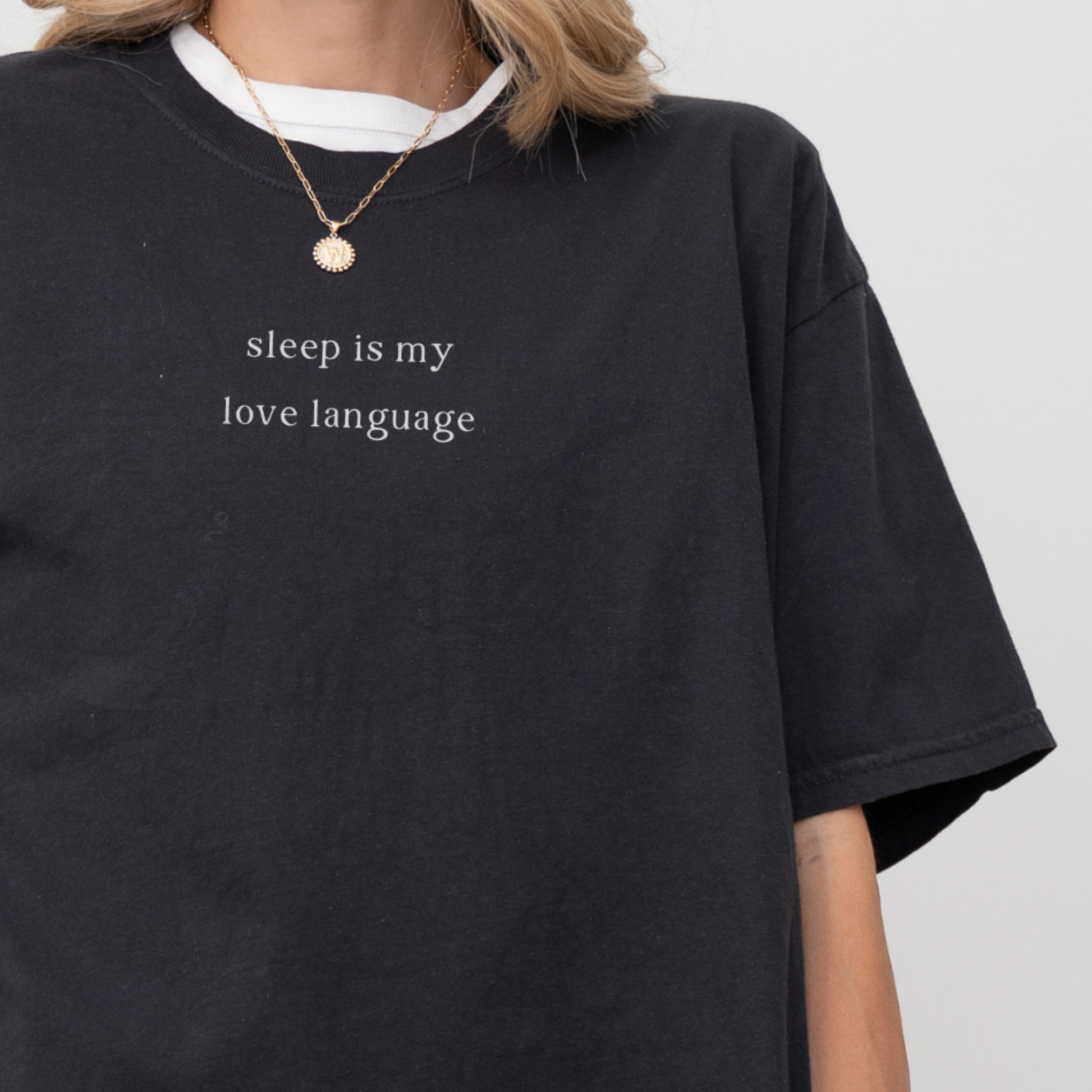 A close up of a woman wearing a black 'sleep is my love language' oversized t-shirt, accessorized with a gold necklace. Minimalist studio product photography for the Barely Sane clothing collection.