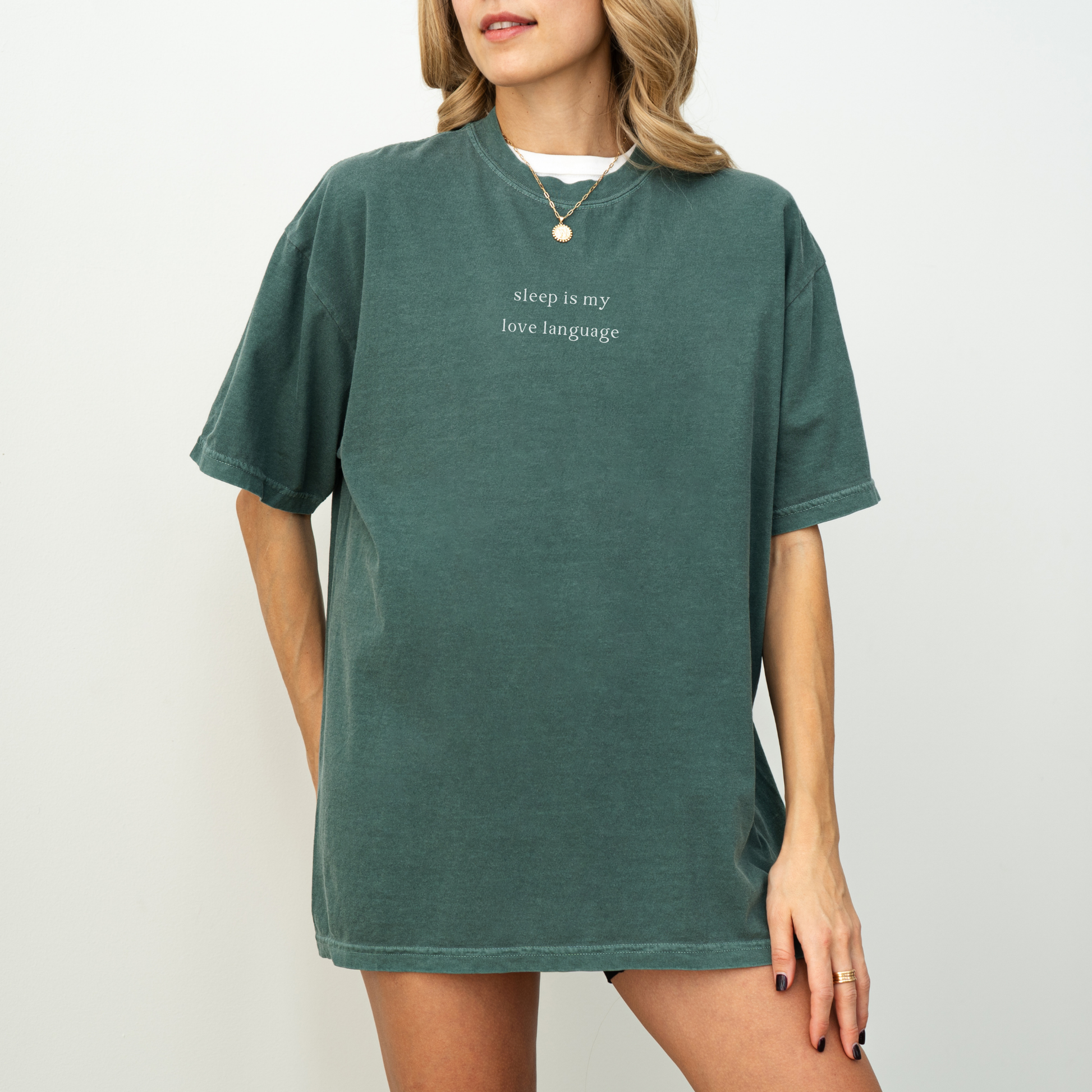 A woman wearing ablue spruce green 'sleep is my love language' oversized t-shirt, accessorized with a gold necklace and a red string bracelet. Minimalist studio product photography for the Barely Sane clothing collection.