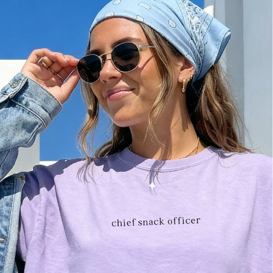 Woman wearing a light purple 'chief snack officer' shirt, denim jacket, and sunglasses outdoors.