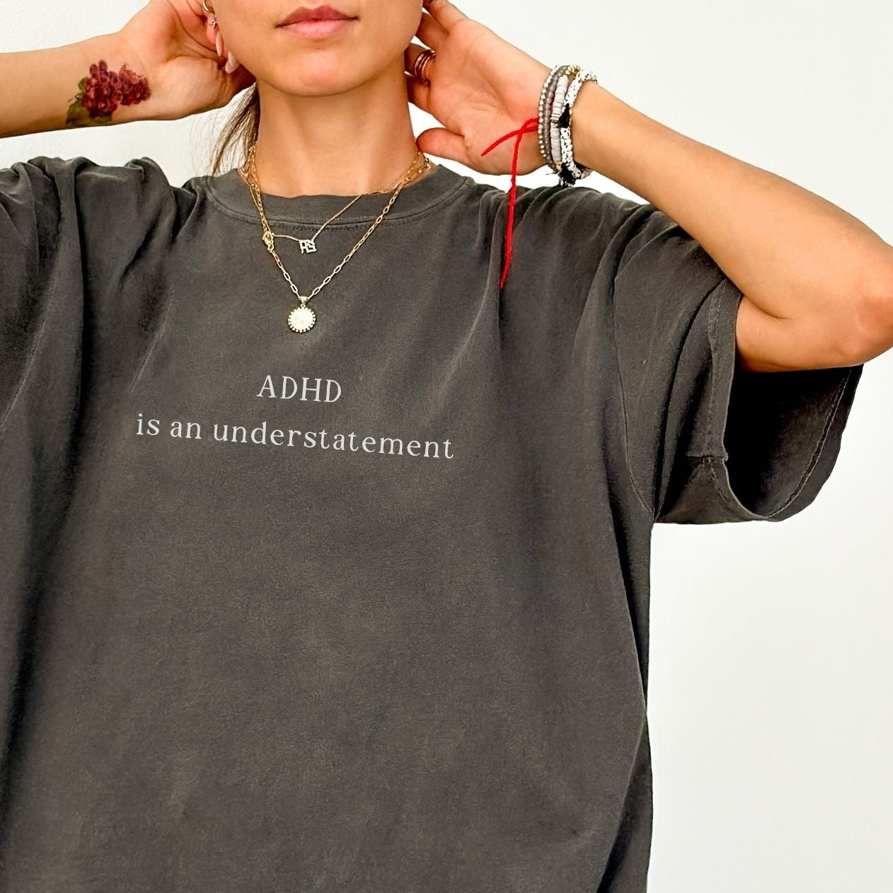 Close-up of an oversized charcoal grey washed t-shirt with white text that reads 'ADHD is an understatement'. Model has hands behind her head, showcasing layered gold necklaces and wrist bracelets against a clean white background.