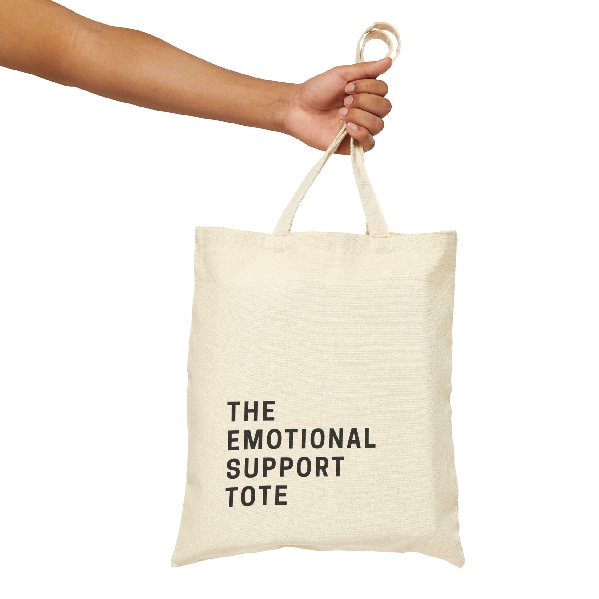 A person's hand holding a natural cotton canvas tote bag. The black text on the bag reads 'THE EMOTIONAL SUPPORT TOTE'. Minimalist product photography on a clean, isolated background by Barely Sane.
