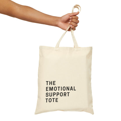 A person's hand holding a natural cotton canvas tote bag. The black text on the bag reads 'THE EMOTIONAL SUPPORT TOTE'. Minimalist product photography on a clean, isolated background by Barely Sane.